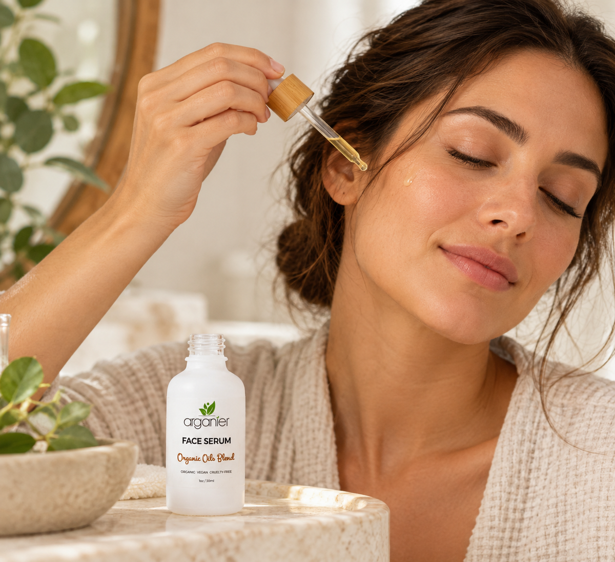 A woman applies a drop of Organic oil Blend face serum to her cheek, with the white and green product bottle resting on a marble surface in the foreground